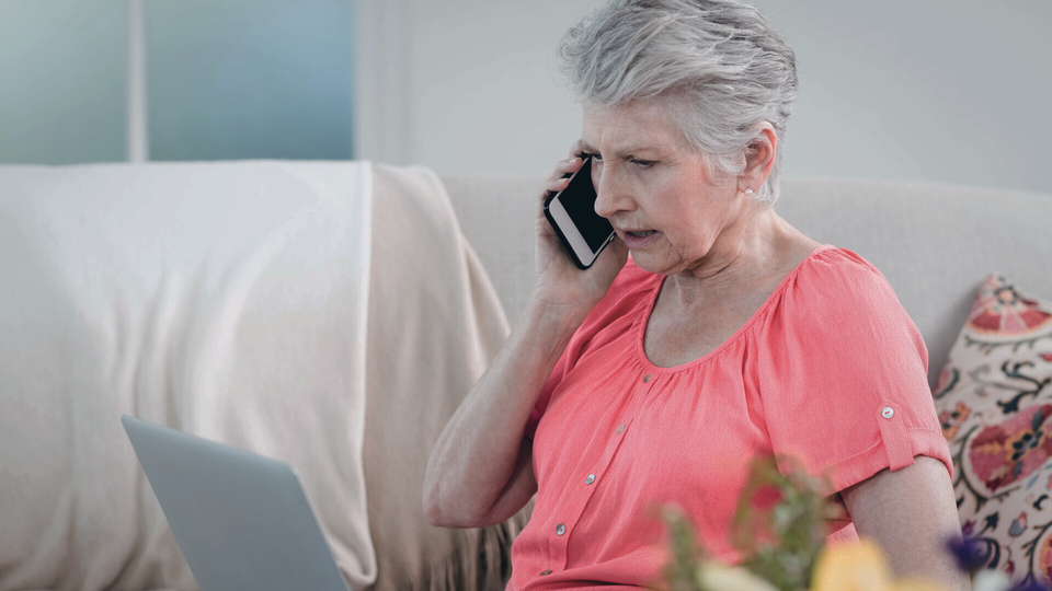 Äldre kvinna pratar i telefon med laptop i knät.