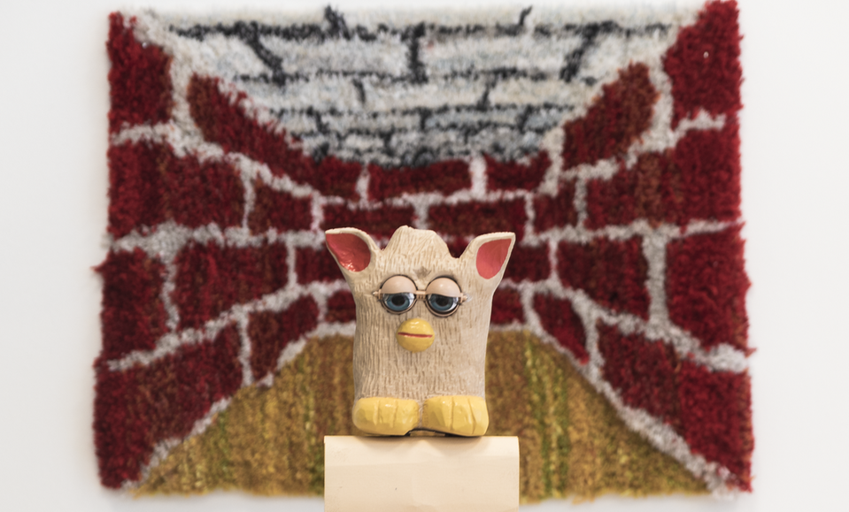 Bilden visar en skulptur av leksaken Furby med stora ögon och öron, näbb och lurvig kropp (nu skulpterad i trä). Furbyn står på en piedestal i trä framför en liten ryamatta som avbildar ett rum i tegel, röda tegelväggar och grå block i taket och gulgrönt golv. Rummet påminner om en scen i ett tv-spel från 90-talet.