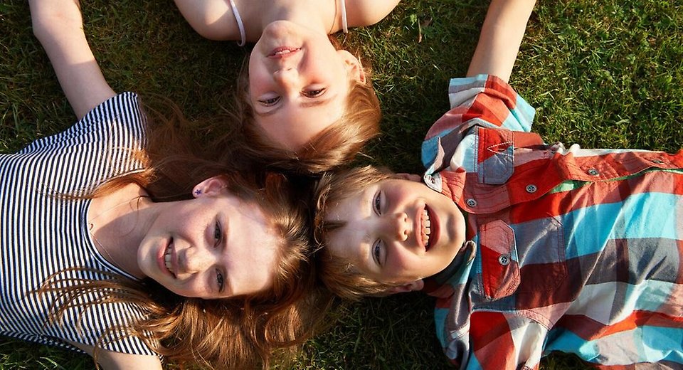 Tre barn ligger i gräset och ler mot kameran.