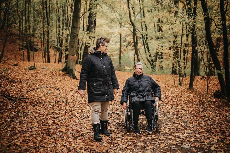 Två glada kvinnor ute i naturen, en kvinna sitter i rullstol 
