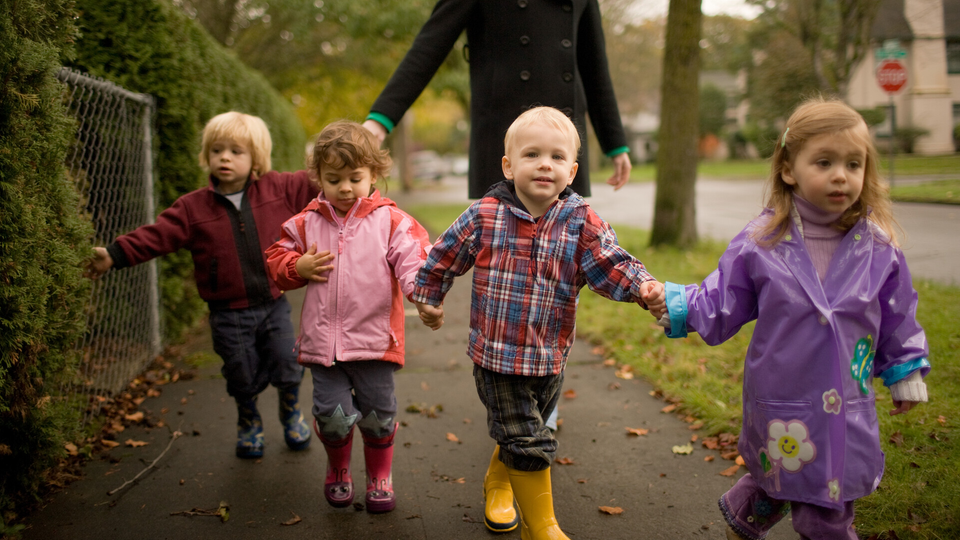 fyra barn går på trottoaren hand i hand, en vuxen person skymtas bakom barnen.